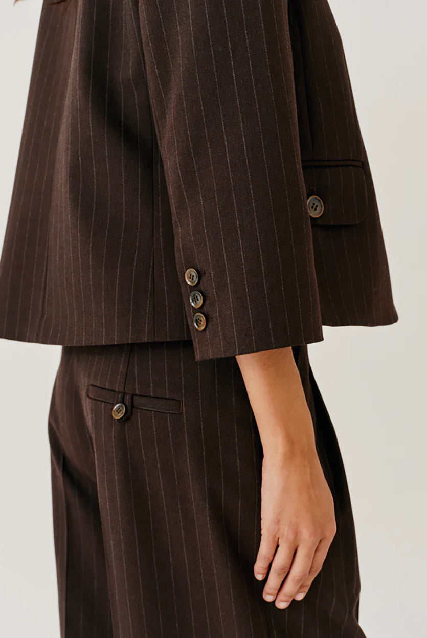 Krótka, pudełkowa marynarka boxy fit Andora Dark Brown z tradycyjnym kołnierzykiem i rękawami długości 3/4. Zapinana na pionowy rząd guzików, przypominających żakiety z lat 90. Marynarka posiada trzy naszywane kieszenie z patkami zapinanymi na guziki- dwie na dole i jedną mniejszą na piersi. Cały materiał zdobiony jest bardzo subtelnym, jasnym, pionowym prążkiem. To klasyczny wzór kojarzony z elegancją biznesową, ale w tym krótkim wydaniu wygląda bardzo modowo.
-Krótki, pudełkowy fason
-Kieszenie na