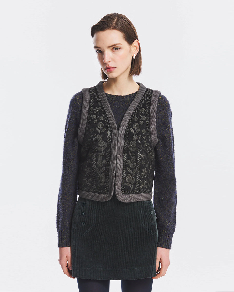 Krótka kamizelka Bertille Black marki Antik Batik o wyraźnym, etnicznym charakterze nawiązującym do stylu boho-chic. Jest to fason sięgający do linii bioder, z otwartym przodem, bez widocznego zapięcia. Przód kamizelki zdobi bogaty, symetryczny haft. Wykonana z mieszanki wełny i bawełny. Wszystkie krawędzie zostały obszyte ozdobną, wzorzystą taśmą z geometrycznym haftem. Zestaw ją z ulubionymi spodniami oraz luźną, białą koszulą z bufiastymi rękawami i wysoką stójką. 