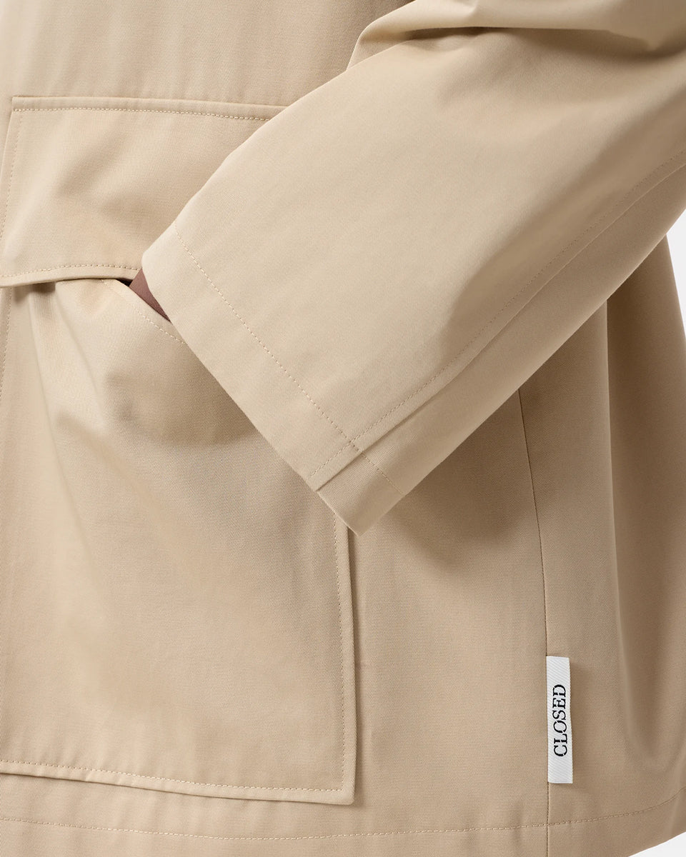 Cienki, krótki, beżowy płaszcz Short coat 97949 CLOSED o luźnym fasonie i prostym kroju. Posiada szerokie, długie rękawy, kołnierzyk i duże, przednie kieszenie z klapkami. Zapinany za pomocą zatrzaskowych guzików