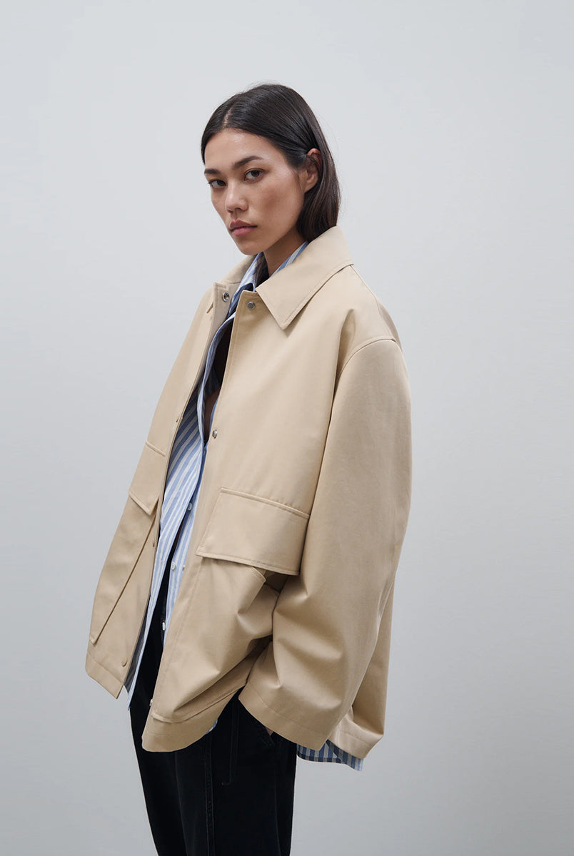 Cienki, krótki, beżowy płaszcz Short coat 97949 CLOSED o luźnym fasonie i prostym kroju. Posiada szerokie, długie rękawy, kołnierzyk i duże, przednie kieszenie z klapkami. Zapinany za pomocą zatrzaskowych guzików
