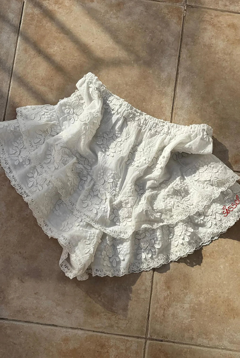 Falbaniaste, kobiece szorty Delphine Off White marki SISSEL EDELBO z elastycznym pasem. Wzdłuż bioder i nogawek ozdobione są falbanami zakończonymi delikatnymi, koronkowymi tasiemkami. Z przodu posiadają haftowany napis 'SISSEL'. Wykonane z bawełny schiffli, zgodnie z ideą marki wykorzystując różnego rodzaju resztki tkanin zamiast produkcji nowych.
-ONE SIZE