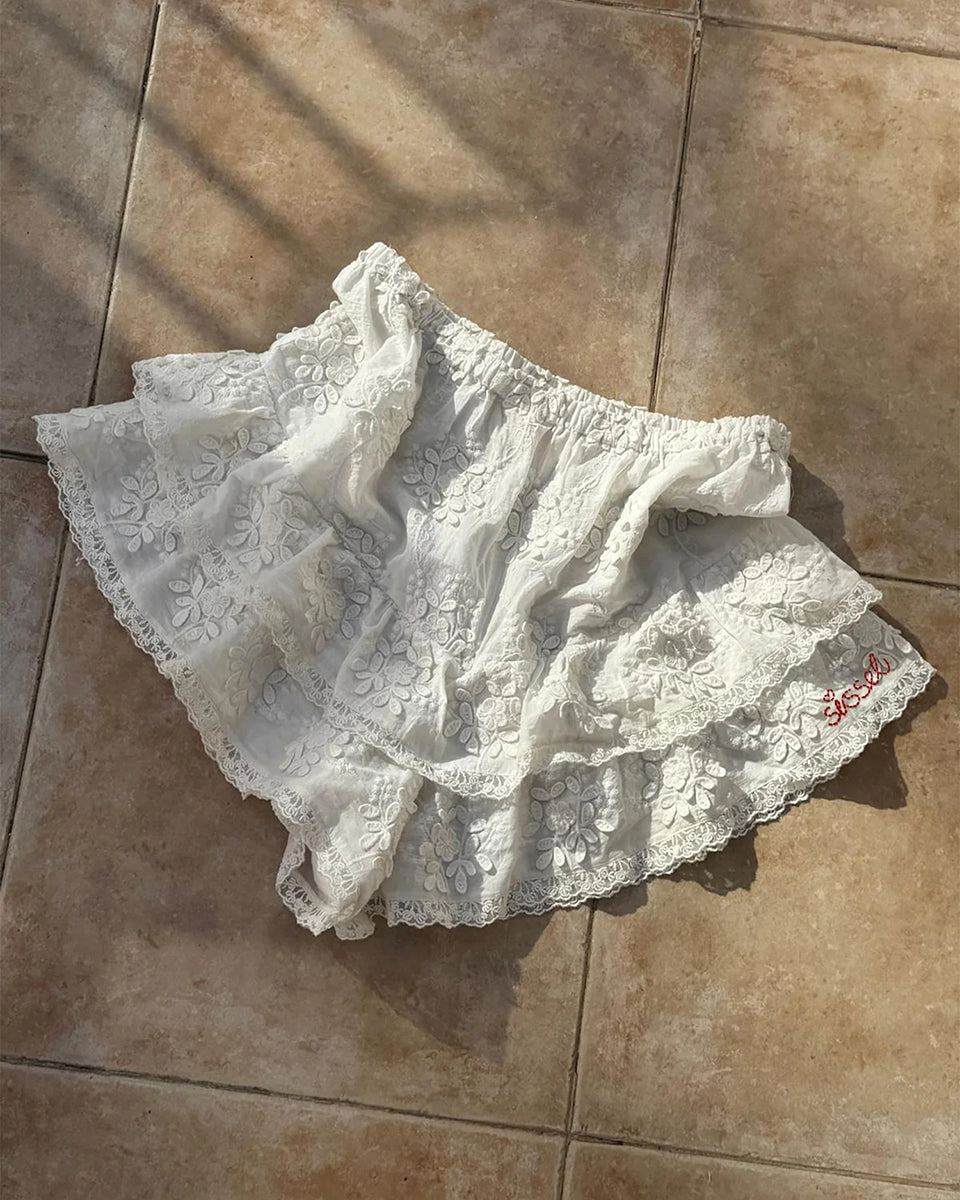 Falbaniaste, kobiece szorty Delphine Off White marki SISSEL EDELBO z elastycznym pasem. Wzdłuż bioder i nogawek ozdobione są falbanami zakończonymi delikatnymi, koronkowymi tasiemkami. Z przodu posiadają haftowany napis 'SISSEL'. Wykonane z bawełny schiffli, zgodnie z ideą marki wykorzystując różnego rodzaju resztki tkanin zamiast produkcji nowych.
-ONE SIZE