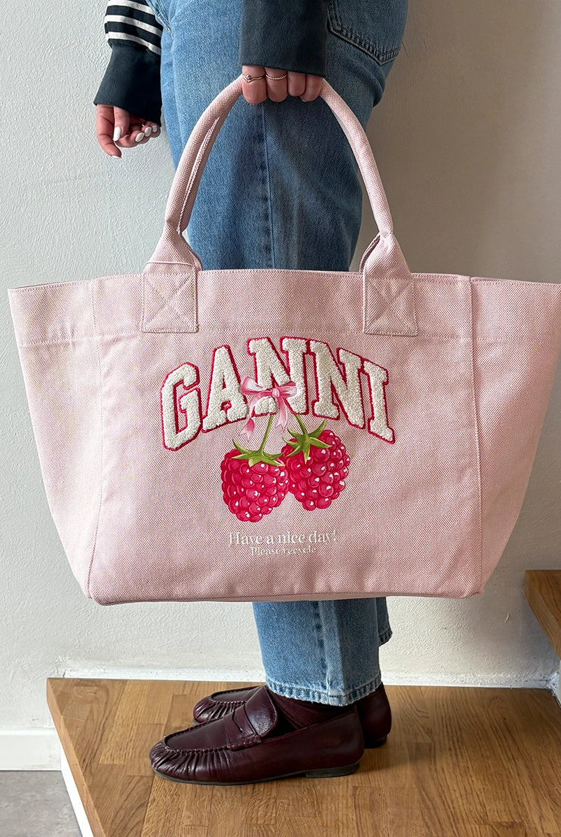 Średniej wielkości, trapezowa, różowa torba shopper 2110 Rasperry GANNI z pluszowym logo marki oraz z uroczym printem z motywem malin. Torba posiada dwie, grube, krótkie rączki do wygodnego trzymania w dłoni oraz odpinany, cienki, długi pasek do wygodnego zawiedzenia na ramieniu. Wewnątrz znajduje się mała kieszonka zapinana na zamek błyskawiczny. Wykonana z grubego, sztywnego, płóciennego materiału. 