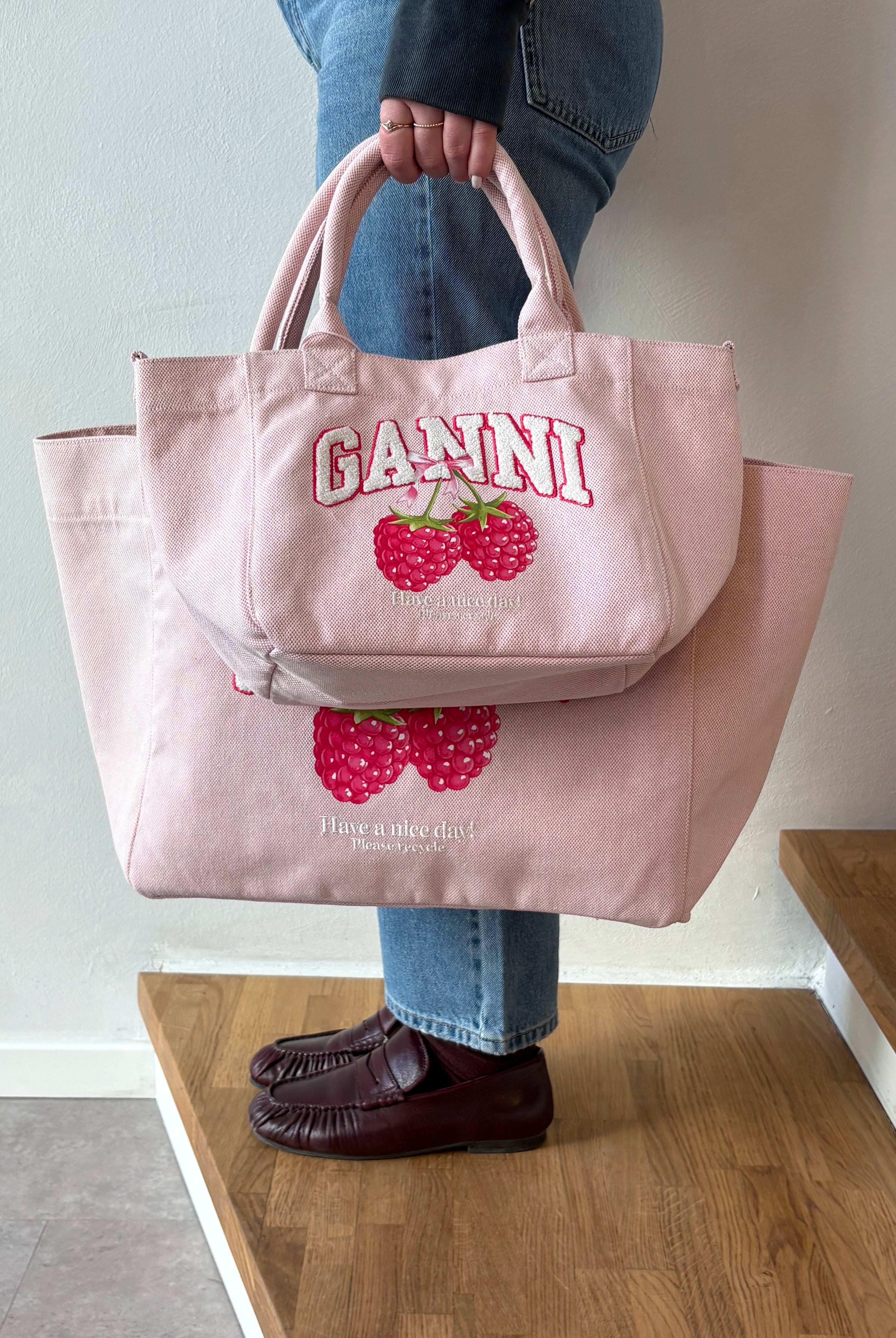 Mała, trapezowa, różowa torba shopper 2110 Rasperry GANNI z pluszowym logo marki oraz z uroczym printem z motywem malin. Torba posiada dwie, grube, krótkie rączki do wygodnego trzymania w dłoni oraz odpinany, cienki, długi pasek do wygodnego zawiedzenia na ramieniu. Wewnątrz znajduje się mała kieszonka zapinana na zamek błyskawiczny. Wykonana z grubego, sztywnego, płóciennego materiału.