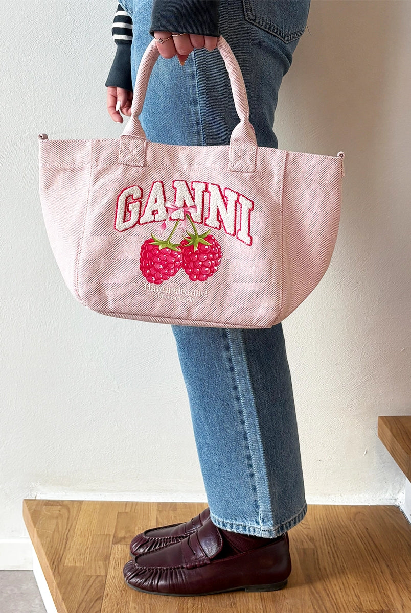 Mała, trapezowa, różowa torba shopper 2110 Rasperry GANNI z pluszowym logo marki oraz z uroczym printem z motywem malin. Torba posiada dwie, grube, krótkie rączki do wygodnego trzymania w dłoni oraz odpinany, cienki, długi pasek do wygodnego zawiedzenia na ramieniu. Wewnątrz znajduje się mała kieszonka zapinana na zamek błyskawiczny. Wykonana z grubego, sztywnego, płóciennego materiału.