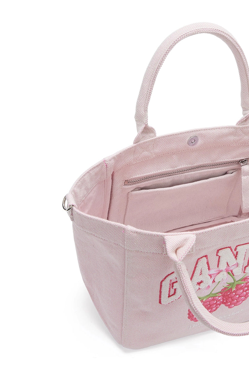 Średniej wielkości, trapezowa, różowa torba shopper 2110 Rasperry GANNI z pluszowym logo marki oraz z uroczym printem z motywem malin. Torba posiada dwie, grube, krótkie rączki do wygodnego trzymania w dłoni oraz odpinany, cienki, długi pasek do wygodnego zawiedzenia na ramieniu. Wewnątrz znajduje się mała kieszonka zapinana na zamek błyskawiczny. Wykonana z grubego, sztywnego, płóciennego materiału. 