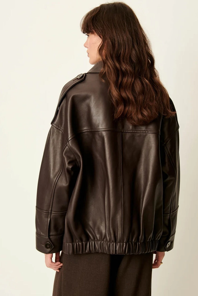 Brązowa, luźna kurtka Harriboyd Coffee Leather francuskiej marki Sessun w stylu klasycznej pilotki. Wykonana jest z gładkiej, ciemnobrązowej skóry o lekkim połysku. Z przodu zapinana jest na duże, czarne guziki, a po bokach ma duże naszywane kieszenie. Rękawy są długie, z mankietami zapinanymi na guzik