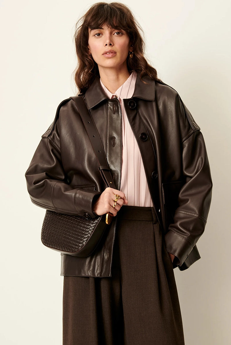 Brązowa, luźna kurtka Harriboyd Coffee Leather francuskiej marki Sessun w stylu klasycznej pilotki. Wykonana jest z gładkiej, ciemnobrązowej skóry o lekkim połysku. Z przodu zapinana jest na duże, czarne guziki, a po bokach ma duże naszywane kieszenie. Rękawy są długie, z mankietami zapinanymi na guzik