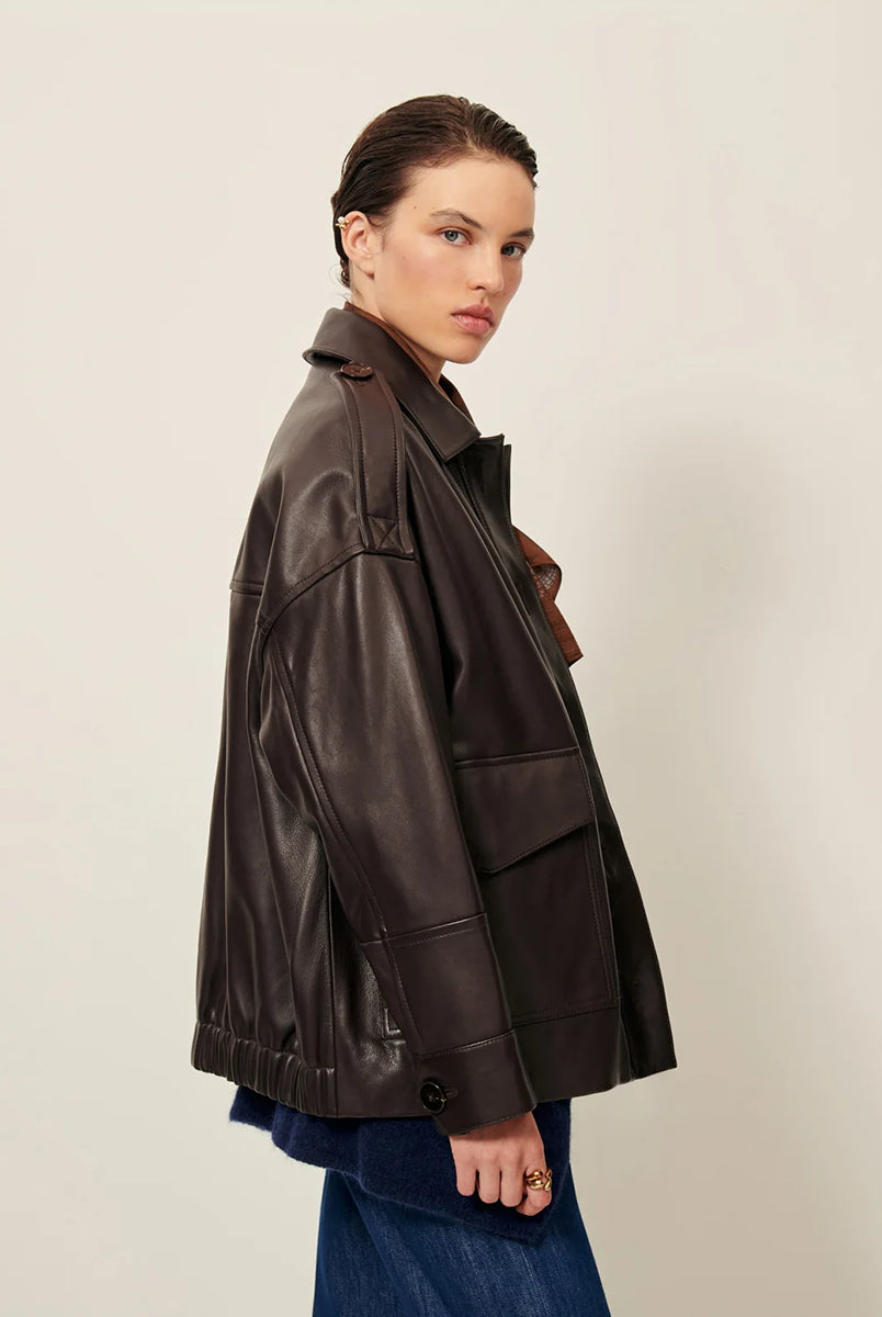 Brązowa, luźna kurtka Harriboyd Coffee Leather francuskiej marki Sessun w stylu klasycznej pilotki. Wykonana jest z gładkiej, ciemnobrązowej skóry o lekkim połysku. Z przodu zapinana jest na duże, czarne guziki, a po bogach ma duże naszywane kieszenie. Rękawy są długie, z mankietami zapinanymi na guzik. Tył kurtki jest prosty, z poziomym szwem przez plecy i elastycznym ściągaczem w dolnej części. 