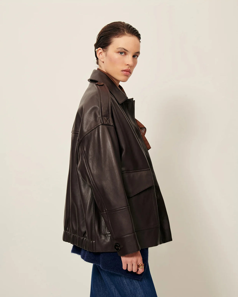 Brązowa, luźna kurtka Harriboyd Coffee Leather francuskiej marki Sessun w stylu klasycznej pilotki. Wykonana jest z gładkiej, ciemnobrązowej skóry o lekkim połysku. Z przodu zapinana jest na duże, czarne guziki, a po bogach ma duże naszywane kieszenie. Rękawy są długie, z mankietami zapinanymi na guzik. Tył kurtki jest prosty, z poziomym szwem przez plecy i elastycznym ściągaczem w dolnej części. 