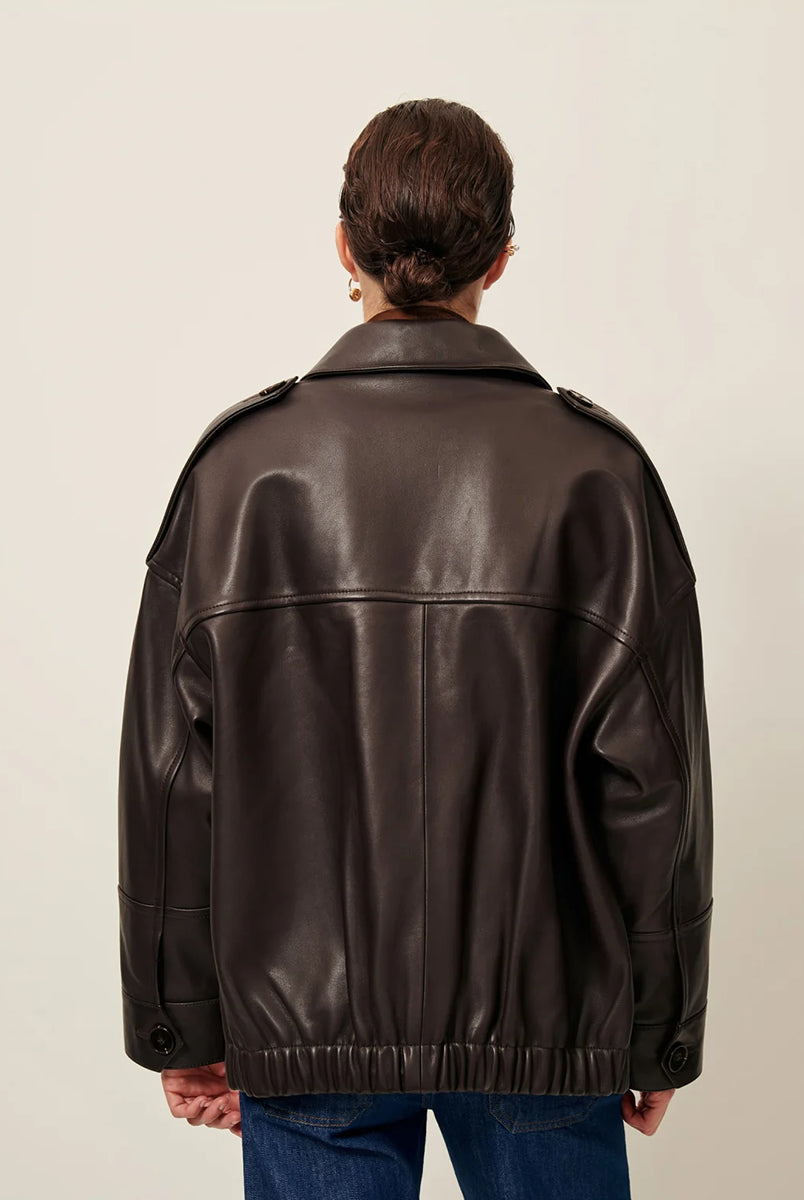 Brązowa, luźna kurtka Harriboyd Coffee Leather francuskiej marki Sessun w stylu klasycznej pilotki. Wykonana jest z gładkiej, ciemnobrązowej skóry o lekkim połysku. Z przodu zapinana jest na duże, czarne guziki, a po bogach ma duże naszywane kieszenie. Rękawy są długie, z mankietami zapinanymi na guzik. Tył kurtki jest prosty, z poziomym szwem przez plecy i elastycznym ściągaczem w dolnej części. 