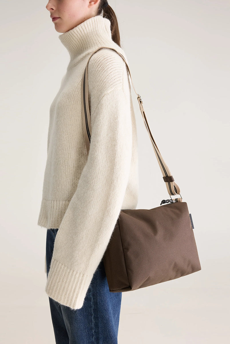 Zapinana na suwak torebka typu cross body Hobbes Dark Roast marki Bellerose z brązowego, matowego nylonu o fakturowanej powierzchni. Wyróżnia się prostokątnym kształtem, miękką konstrukcją oraz regulowanym paskiem w paski i silikonowym logo. 