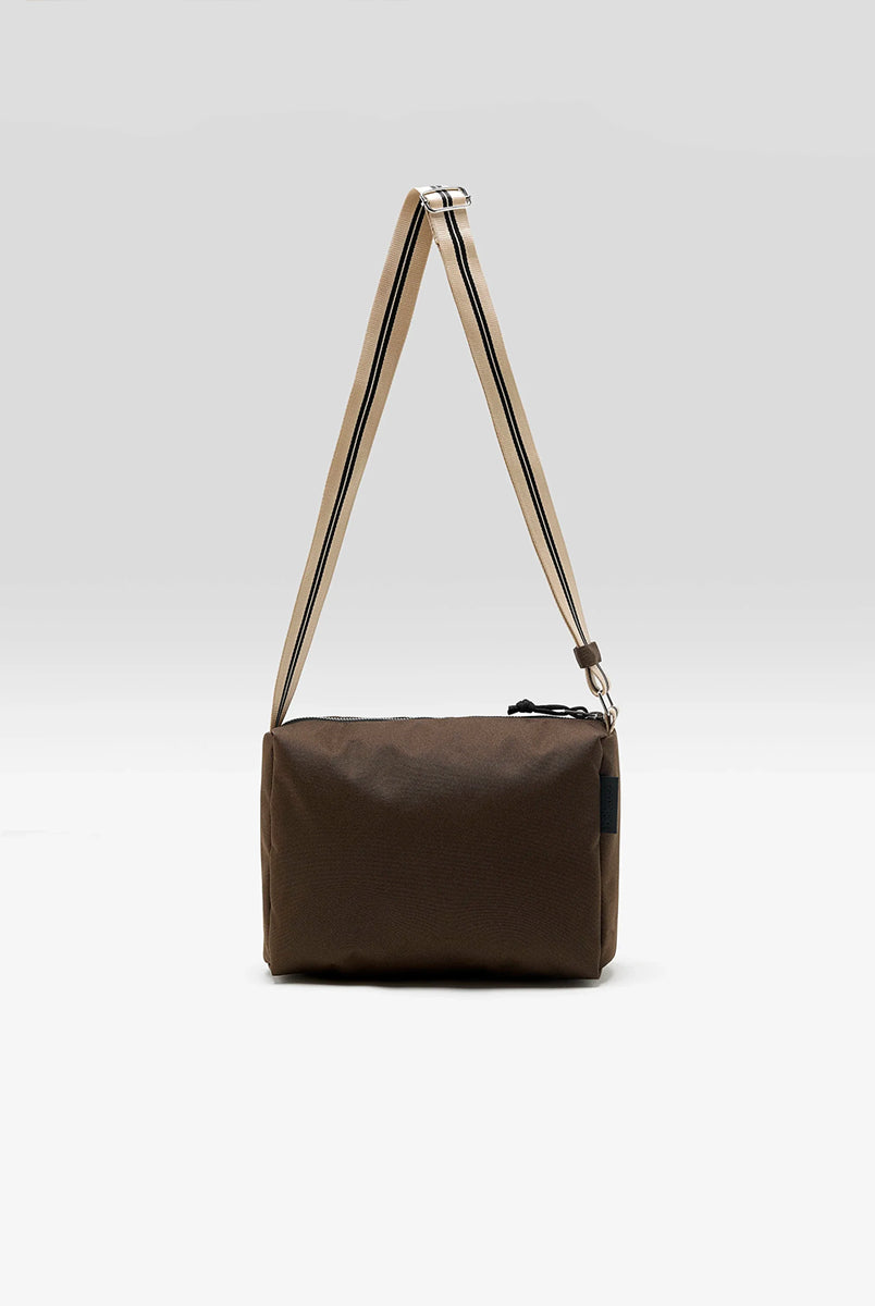 Zapinana na suwak torebka typu cross body Hobbes Dark Roast marki Bellerose z brązowego, matowego nylonu o fakturowanej powierzchni. Wyróżnia się prostokątnym kształtem, miękką konstrukcją oraz regulowanym paskiem w paski i silikonowym logo. 