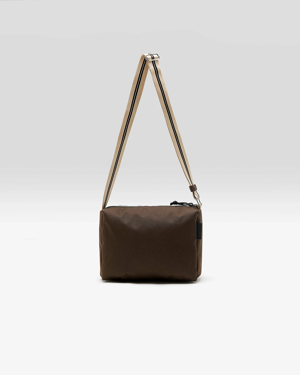Zapinana na suwak torebka typu cross body Hobbes Dark Roast marki Bellerose z brązowego, matowego nylonu o fakturowanej powierzchni. Wyróżnia się prostokątnym kształtem, miękką konstrukcją oraz regulowanym paskiem w paski i silikonowym logo. 