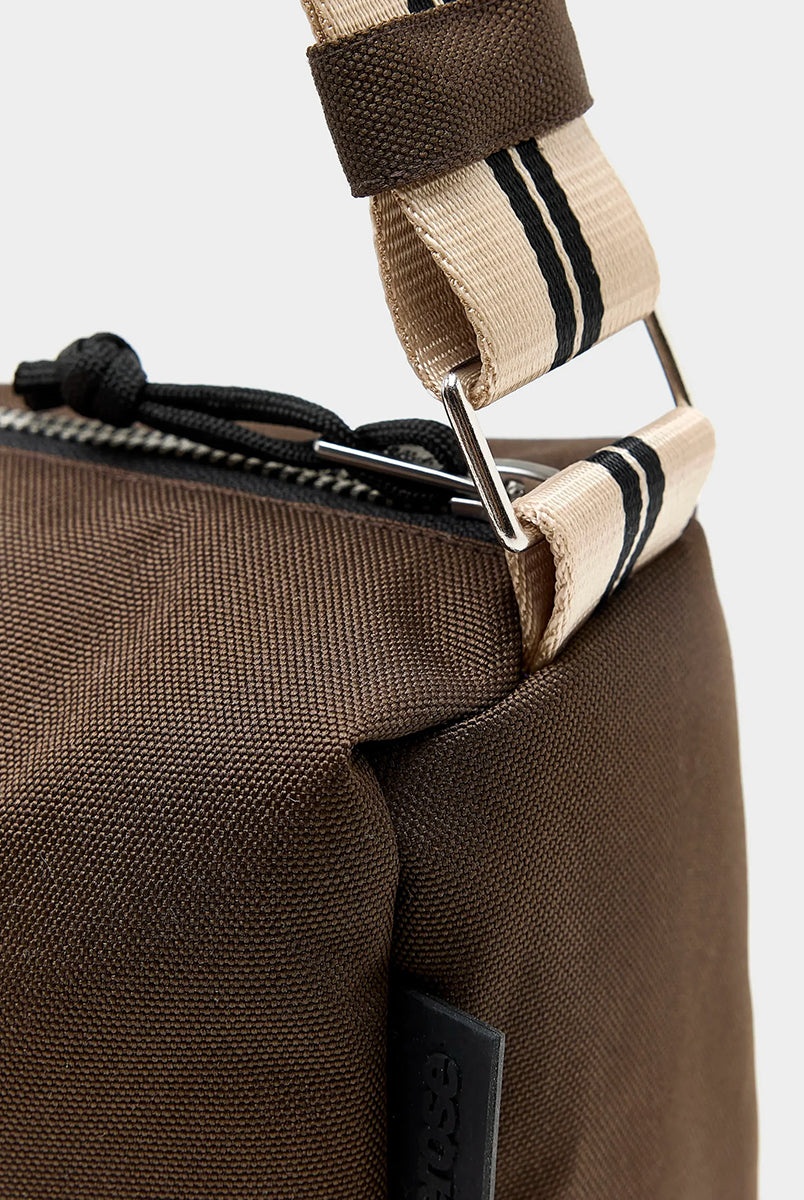 Zapinana na suwak torebka typu cross body Hobbes Dark Roast marki Bellerose z brązowego, matowego nylonu o fakturowanej powierzchni. Wyróżnia się prostokątnym kształtem, miękką konstrukcją oraz regulowanym paskiem w paski i silikonowym logo. 