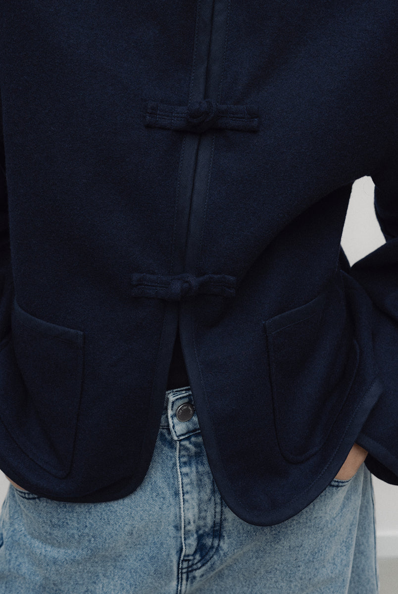 Elegancka, ciemnogranatowa, krótka kurtka Indie Black Iris o fasonie inspirowanym stylem wschodnim. Model ma prosty krój z okrągłym dekoltem bez kołnierzyka oraz długie rękawy o swobodnej linii. Charakterystycznym elementem są ozdobne, tekstylne zapięcia typu chińskiego, zwane węzełkami, które biegną wzdłuż przedniej plisy. Kurtka posiada również dwie duże, naszywane kieszenie oraz zaokrąglone doły po bokach, co nadaje jej miękki, nowoczesny wygląd. Wykonana z mieszanki z wełną.
-Inspirowana stylem wschodn
