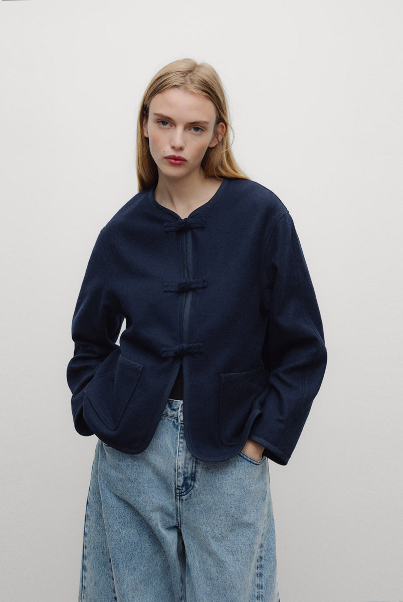Elegancka, ciemnogranatowa, krótka kurtka Indie Black Iris o fasonie inspirowanym stylem wschodnim. Model ma prosty krój z okrągłym dekoltem bez kołnierzyka oraz długie rękawy o swobodnej linii. Charakterystycznym elementem są ozdobne, tekstylne zapięcia typu chińskiego, zwane węzełkami, które biegną wzdłuż przedniej plisy. Kurtka posiada również dwie duże, naszywane kieszenie oraz zaokrąglone doły po bokach, co nadaje jej miękki, nowoczesny wygląd. Wykonana z mieszanki z wełną.
-Inspirowana stylem wschodn