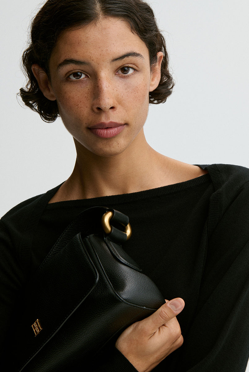 Minimalistyczna, elegancka torebka Lellie Black By Malene Birger o prostokątnym kształcie z lekko zaokrąglonymi bokami. Zapinana na zatrzask magnetyczny. Posiada krótki, szeroki pasek na ramię, który ozdobiony jest złotymi elementami. Na środku klapy umieszczono metalowy emblemat marki w kolorze złotym. W środku znajduje się mała kieszonka na drobiazgi, a całość wykończona jest gładką podszewką. Wykonana w 100% ze skóry. 