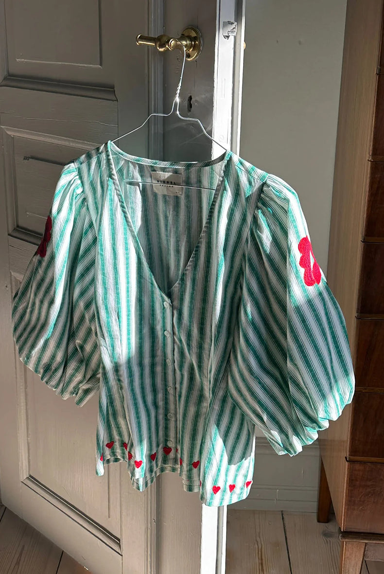 Kobieca bluzka koszulowa Lotta Green Stripes SISSEL EDELBO z dopasowaną talią i marszczeniem z tyłu. Ma głęboki dekolt w serek i zapięcie z przodu na guziki z masy perłowej. Rękawy mają długość 3/4 i zakończone są gumką. Ozdobiona jest delikatnym haftem kwiatowym i serduszkami. Wykonana z organicznej bawełny popelinowej w paski.
-Dopasowana talia
-100% bawełna organiczna
-Zamów bluzkę w swoim standardowym rozmiarze 