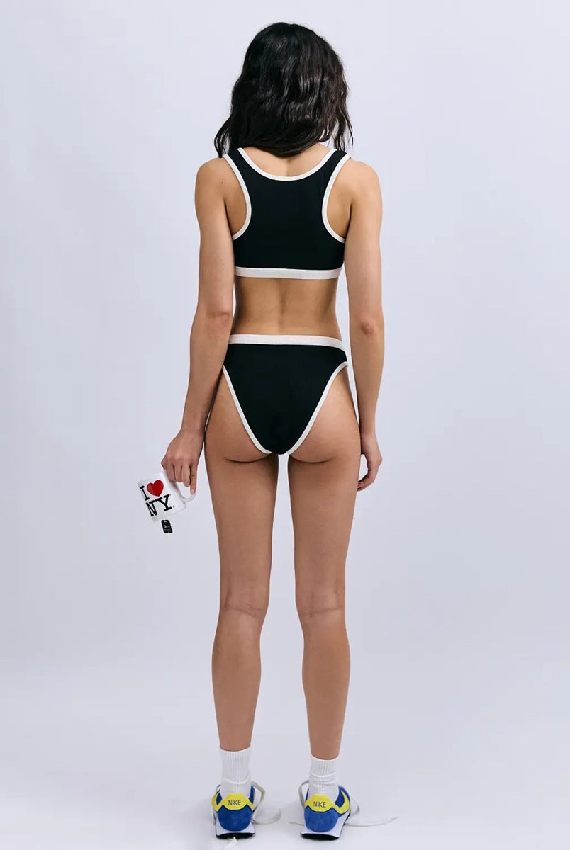 Miękki sportowy stanik top Rika Love Love Stories na grubych ramiączkach. Wykonany z miękkiej tkaniny z prążkowaną fakturą i białą, grubą lamówką. Na gumie pod biustem posiada niewielką naszywkę z logo marki. 