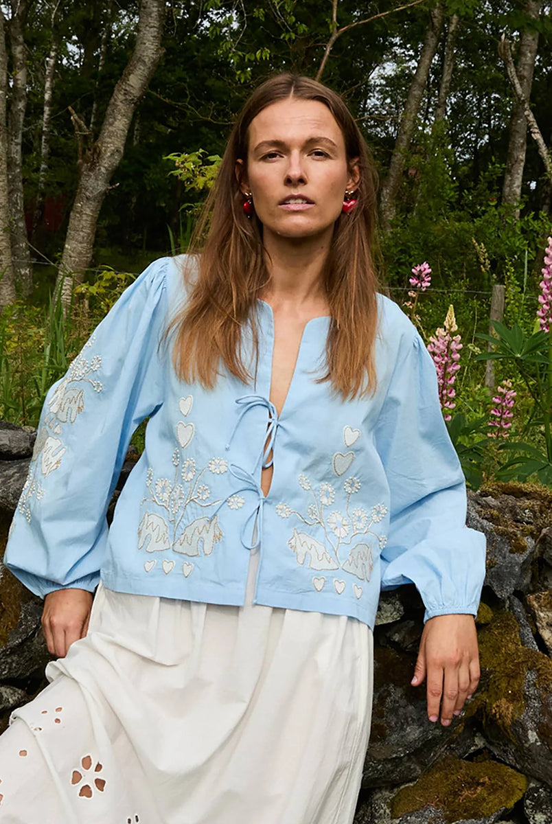 Luźna, krótka bluzka Maggan Light Blue SISSEL EDELBO z szerokimi, obszernymi rękawami z marszczeniami na ramionach. Rękawy zakończone są gumką. Dekolt jest delikatnie wyprofilowany i wiązany z przodu na dwa sznurki. Bluzkę zdobi haft z delikatnymi koralikami. 
