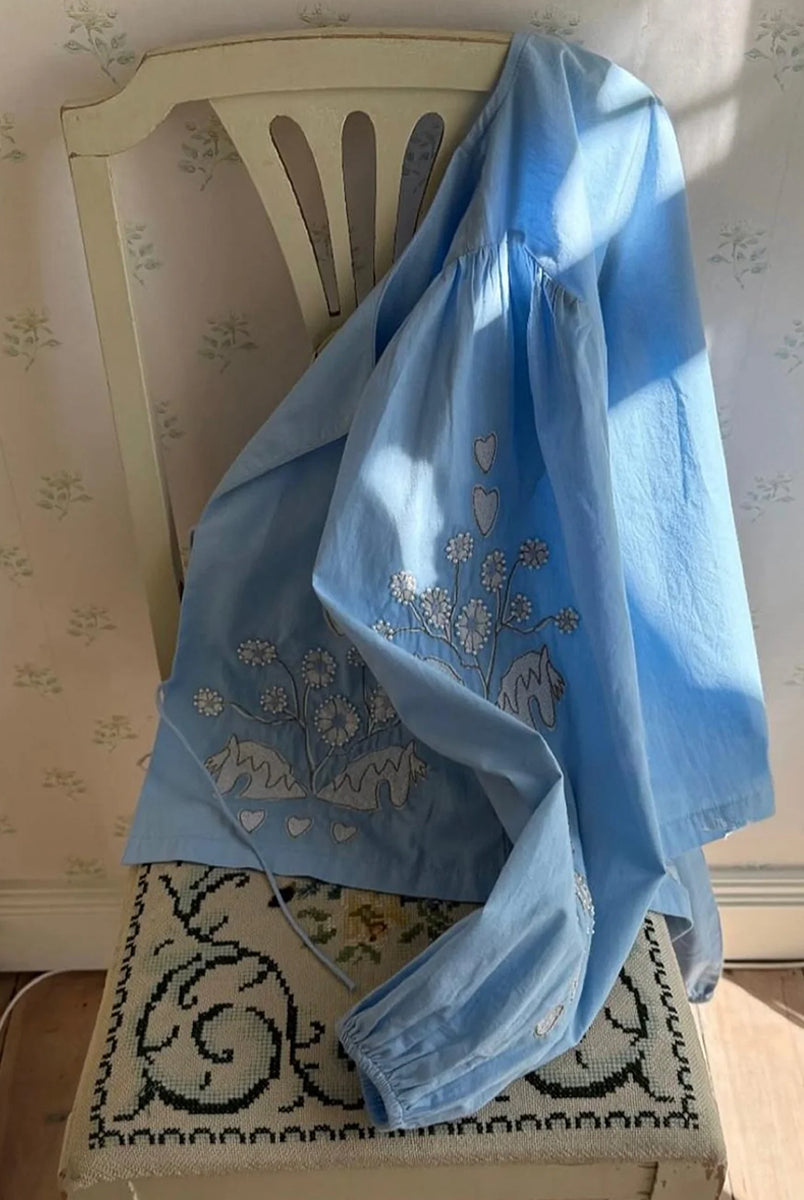 Luźna, krótka bluzka Maggan Light Blue SISSEL EDELBO z szerokimi, obszernymi rękawami z marszczeniami na ramionach. Rękawy zakończone są gumką. Dekolt jest delikatnie wyprofilowany i wiązany z przodu na dwa sznurki. Bluzkę zdobi haft z delikatnymi koralikami. 