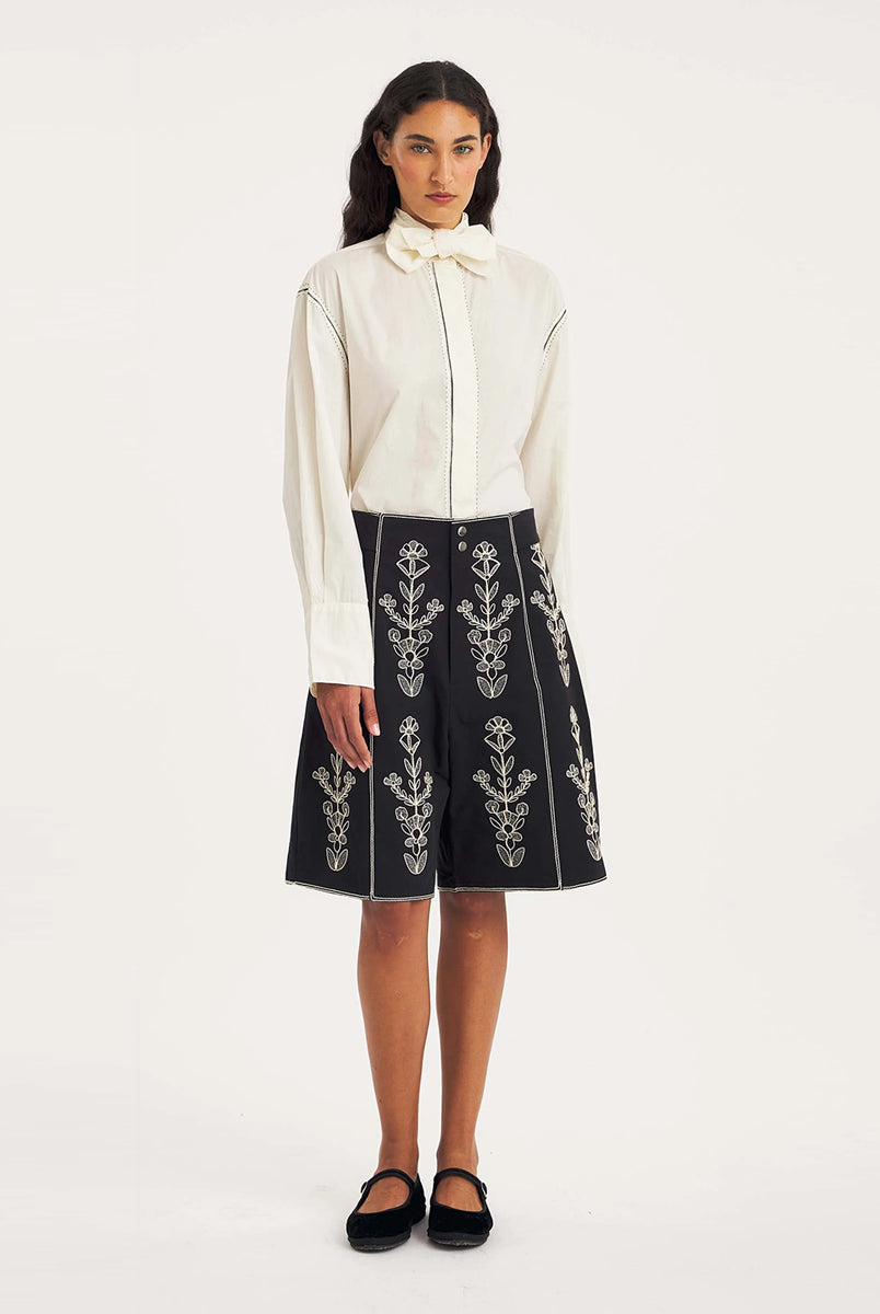 Dłuższe, eleganckie szorty typu bermudy Maria Black marki Antik Batik sięgające do kolan. Mają luźne, szerokie i prosto ścięte nogawki oraz wysoki stan z szerokim pasem i szlufkami. Zapinane są z przodu na dwa jasne guziki umieszczone pionowo. Wzdłuż nogawek oraz wokół kieszeni i paska mają kontrastowe, jasne lamówki. Przód, jak i tył zdobią bogate białe hafty o motywie roślinnym. Hafty umieszczone są symetrycznie w pionowych rzędach, co nawiązuje do tradycyjnego stylu boho.
-Luźne, proste nogawki
-