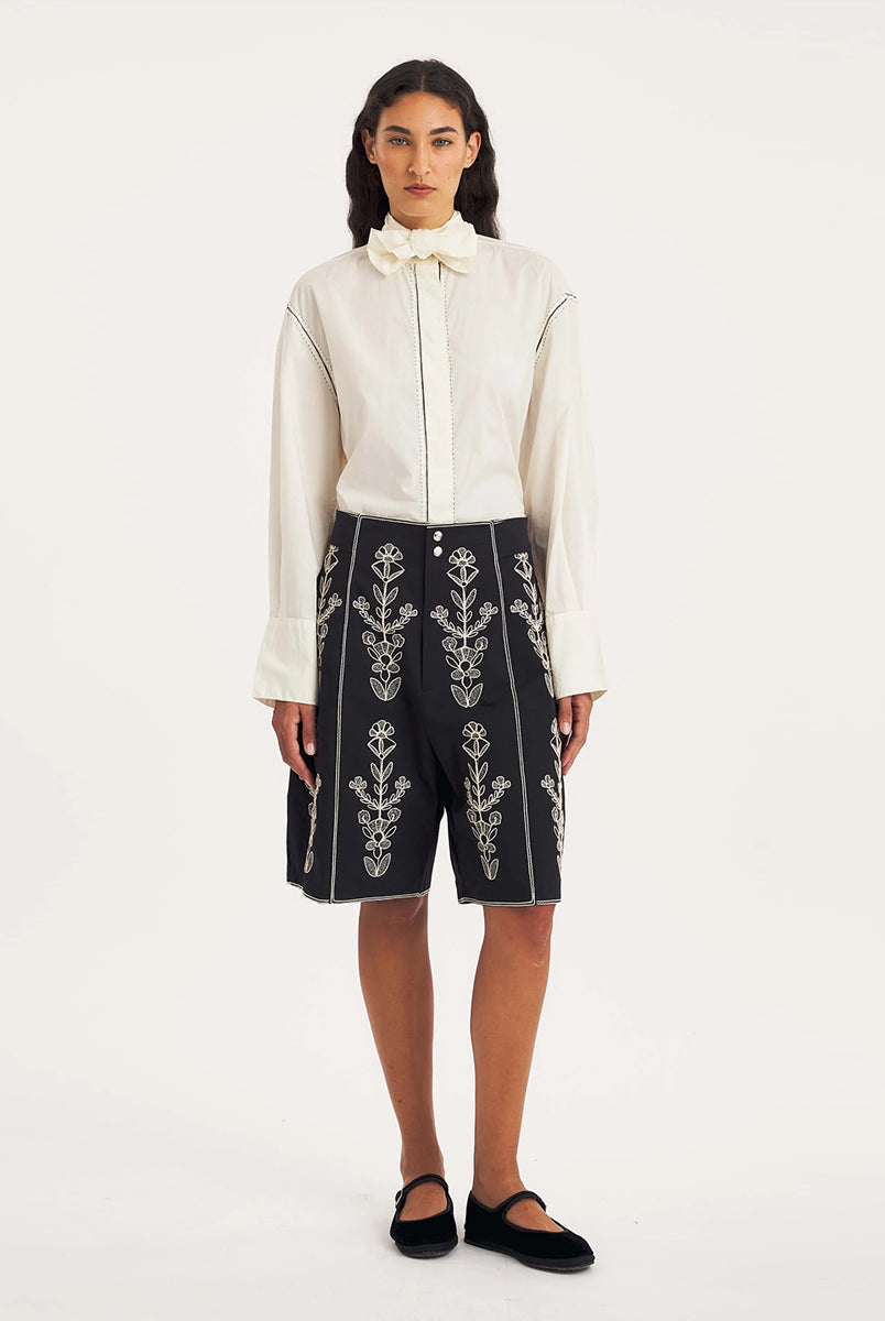 Dłuższe, eleganckie szorty typu bermudy Maria Black marki Antik Batik sięgające do kolan. Mają luźne, szerokie i prosto ścięte nogawki oraz wysoki stan z szerokim pasem i szlufkami. Zapinane są z przodu na dwa jasne guziki umieszczone pionowo. Wzdłuż nogawek oraz wokół kieszeni i paska mają kontrastowe, jasne lamówki. Przód, jak i tył zdobią bogate białe hafty o motywie roślinnym. Hafty umieszczone są symetrycznie w pionowych rzędach, co nawiązuje do tradycyjnego stylu boho.
-Luźne, proste nogawki
-