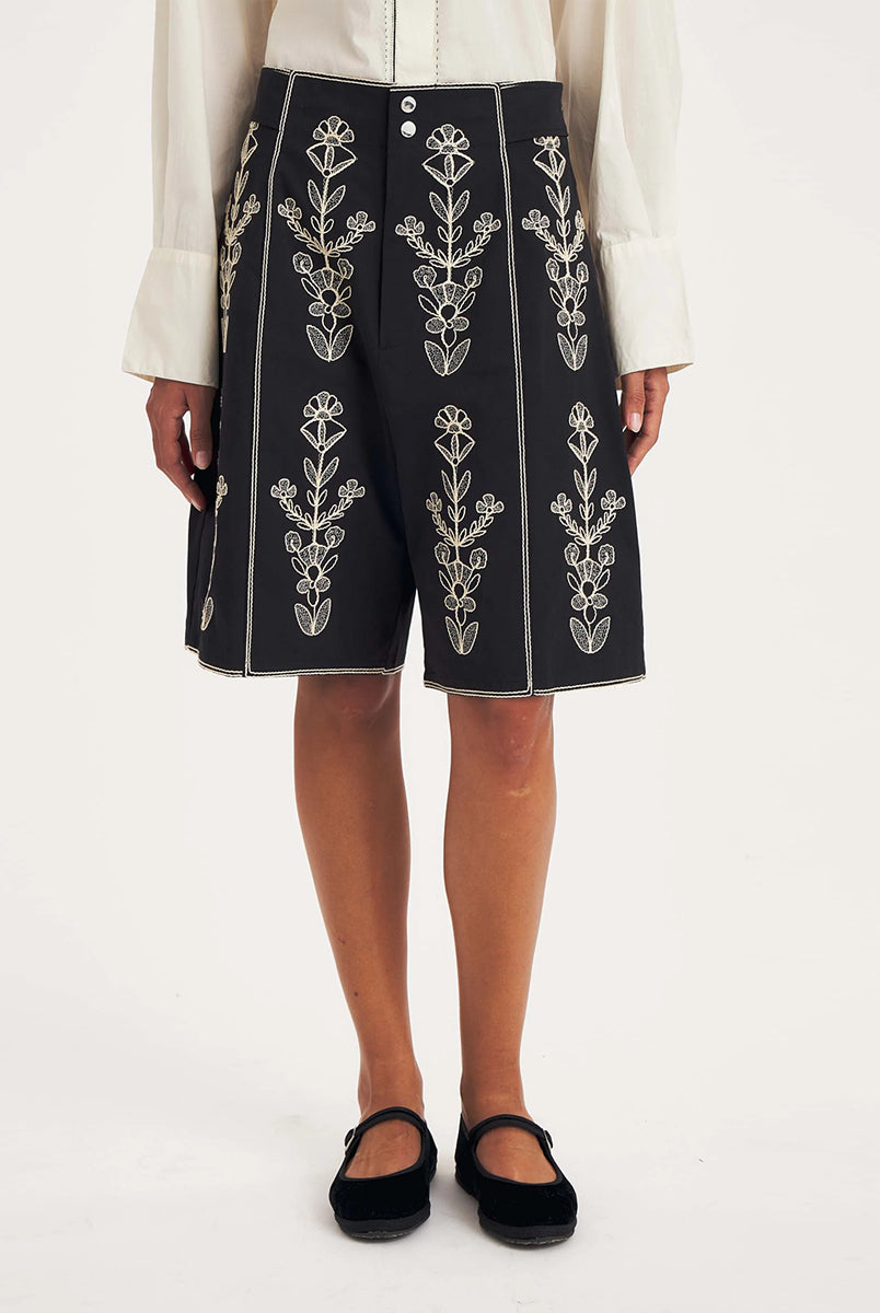 Dłuższe, eleganckie szorty typu bermudy Maria Black marki Antik Batik sięgające do kolan. Mają luźne, szerokie i prosto ścięte nogawki oraz wysoki stan z szerokim pasem i szlufkami. Zapinane są z przodu na dwa jasne guziki umieszczone pionowo. Wzdłuż nogawek oraz wokół kieszeni i paska mają kontrastowe, jasne lamówki. Przód, jak i tył zdobią bogate białe hafty o motywie roślinnym. Hafty umieszczone są symetrycznie w pionowych rzędach, co nawiązuje do tradycyjnego stylu boho.
-Luźne, proste nogawki
-