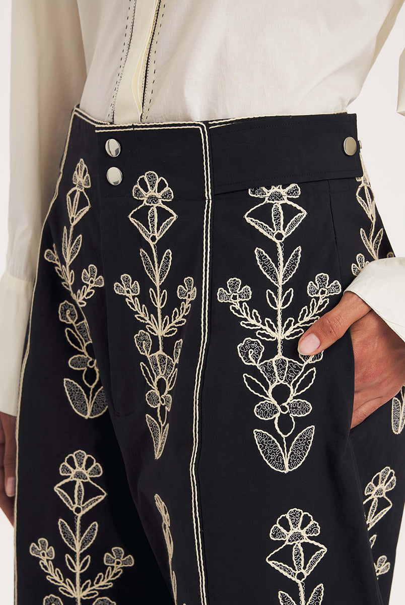 Dłuższe, eleganckie szorty typu bermudy Maria Black marki Antik Batik sięgające do kolan. Mają luźne, szerokie i prosto ścięte nogawki oraz wysoki stan z szerokim pasem i szlufkami. Zapinane są z przodu na dwa jasne guziki umieszczone pionowo. Wzdłuż nogawek oraz wokół kieszeni i paska mają kontrastowe, jasne lamówki. Przód, jak i tył zdobią bogate białe hafty o motywie roślinnym. Hafty umieszczone są symetrycznie w pionowych rzędach, co nawiązuje do tradycyjnego stylu boho.
-Luźne, proste nogawki
-