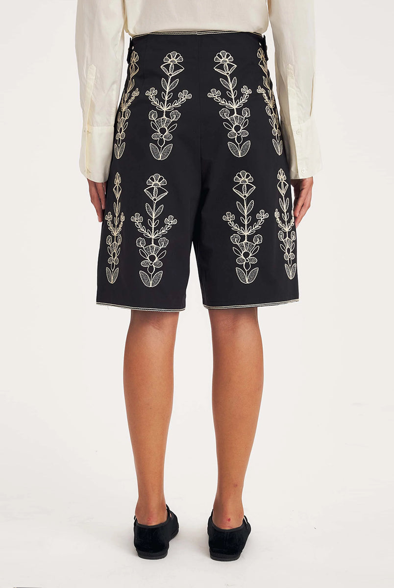 Dłuższe, eleganckie szorty typu bermudy Maria Black marki Antik Batik sięgające do kolan. Mają luźne, szerokie i prosto ścięte nogawki oraz wysoki stan z szerokim pasem i szlufkami. Zapinane są z przodu na dwa jasne guziki umieszczone pionowo. Wzdłuż nogawek oraz wokół kieszeni i paska mają kontrastowe, jasne lamówki. Przód, jak i tył zdobią bogate białe hafty o motywie roślinnym. Hafty umieszczone są symetrycznie w pionowych rzędach, co nawiązuje do tradycyjnego stylu boho.
-Luźne, proste nogawki
-