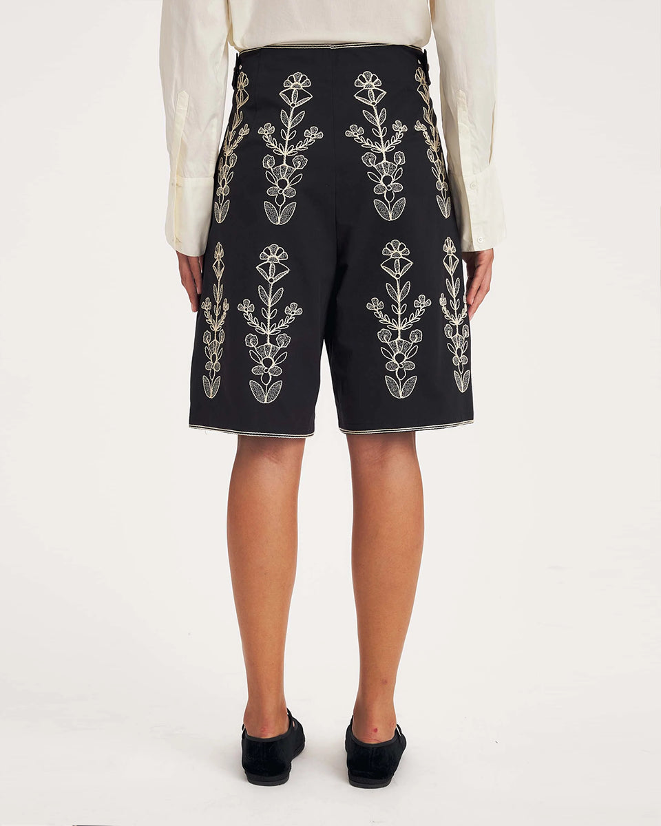 Dłuższe, eleganckie szorty typu bermudy Maria Black marki Antik Batik sięgające do kolan. Mają luźne, szerokie i prosto ścięte nogawki oraz wysoki stan z szerokim pasem i szlufkami. Zapinane są z przodu na dwa jasne guziki umieszczone pionowo. Wzdłuż nogawek oraz wokół kieszeni i paska mają kontrastowe, jasne lamówki. Przód, jak i tył zdobią bogate białe hafty o motywie roślinnym. Hafty umieszczone są symetrycznie w pionowych rzędach, co nawiązuje do tradycyjnego stylu boho.
-Luźne, proste nogawki
-