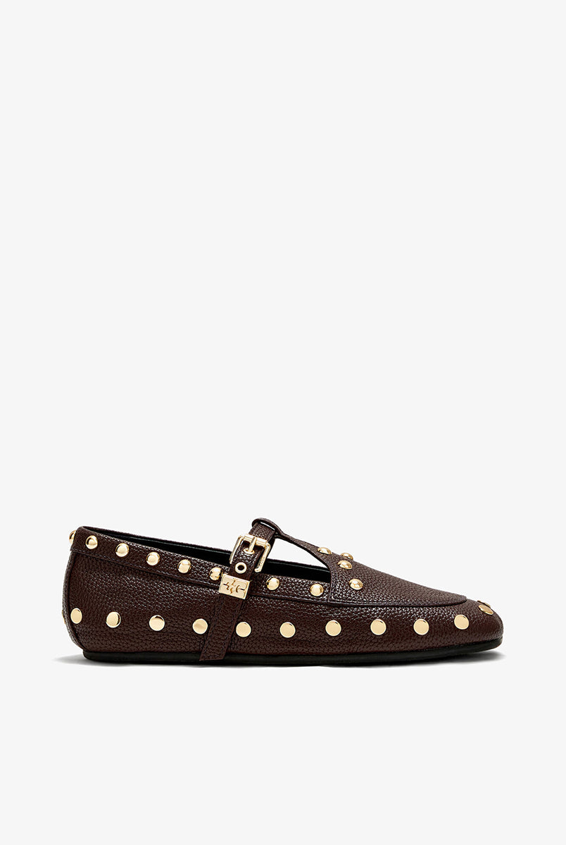 Płaskie, ciemnobrązowe mokasyny Mary Jenny Studs GANNI Chicory Coffee o zaokrąglonych noskach. Cholewkę zdobią liczne, gęsto rozmieszczone złote piny (ćwieki). Posiadają regulowane paski zapinane na złotą klamrę zakończoną charakterystycznym logo GANNI Butterfly po zewnętrznej stronie stopy. Buty mają wyraźną, groszkowaną fakturę. Rozmiarówka jest standardowa, a wysokość obcasa to 0,7 cm.
-Standardowa rozmiarówka