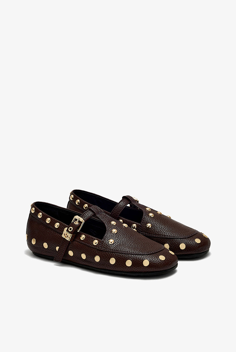 Płaskie, ciemnobrązowe mokasyny Mary Jenny Studs GANNI Chicory Coffee o zaokrąglonych noskach. Cholewkę zdobią liczne, gęsto rozmieszczone złote piny (ćwieki). Posiadają regulowane paski zapinane na złotą klamrę zakończoną charakterystycznym logo GANNI Butterfly po zewnętrznej stronie stopy. Buty mają wyraźną, groszkowaną fakturę. Rozmiarówka jest standardowa, a wysokość obcasa to 0,7 cm.
-Standardowa rozmiarówka