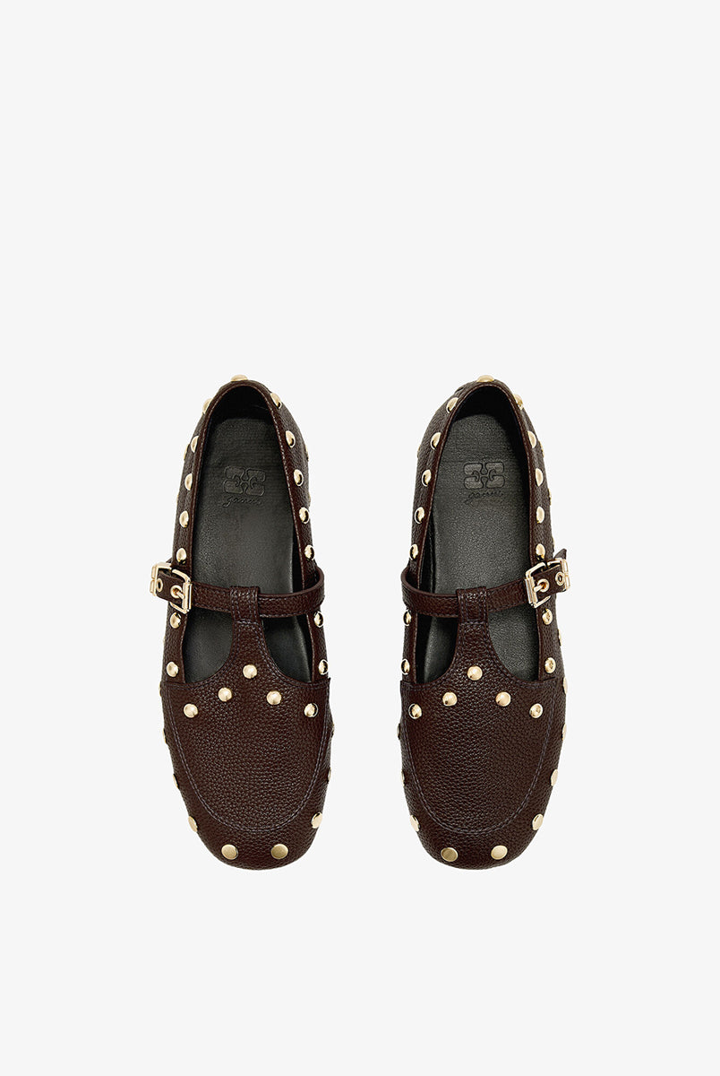 Płaskie, ciemnobrązowe mokasyny Mary Jenny Studs GANNI Chicory Coffee o zaokrąglonych noskach. Cholewkę zdobią liczne, gęsto rozmieszczone złote piny (ćwieki). Posiadają regulowane paski zapinane na złotą klamrę zakończoną charakterystycznym logo GANNI Butterfly po zewnętrznej stronie stopy. Buty mają wyraźną, groszkowaną fakturę. Rozmiarówka jest standardowa, a wysokość obcasa to 0,7 cm.
-Standardowa rozmiarówka