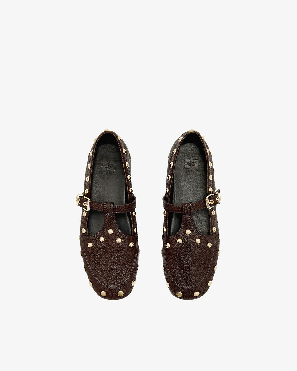 Płaskie, ciemnobrązowe mokasyny Mary Jenny Studs GANNI Chicory Coffee o zaokrąglonych noskach. Cholewkę zdobią liczne, gęsto rozmieszczone złote piny (ćwieki). Posiadają regulowane paski zapinane na złotą klamrę zakończoną charakterystycznym logo GANNI Butterfly po zewnętrznej stronie stopy. Buty mają wyraźną, groszkowaną fakturę. Rozmiarówka jest standardowa, a wysokość obcasa to 0,7 cm.
-Standardowa rozmiarówka