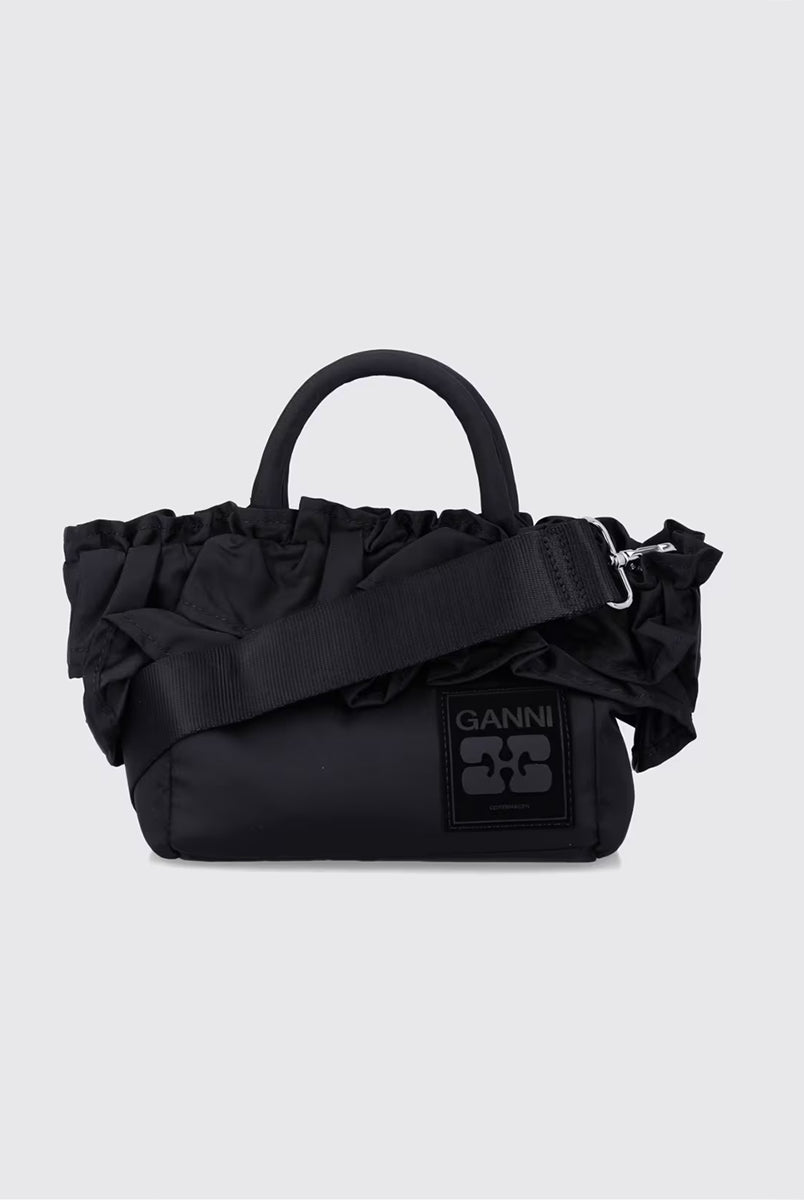 Torba typu tote 7187 Mini Shopper Black marki GANNI z uchwytami i paskiem na ramię. Posiada jedną dużą komorę z wewnętrzną kieszenią zapinaną na zamek. U dołu znajduje się niewielkie logo marki. Wyjątkowości dodaje zdobienie w postaci falbanek. Zaprojektowana, aby pomieścić najpotrzebniejsze drobiazgi.