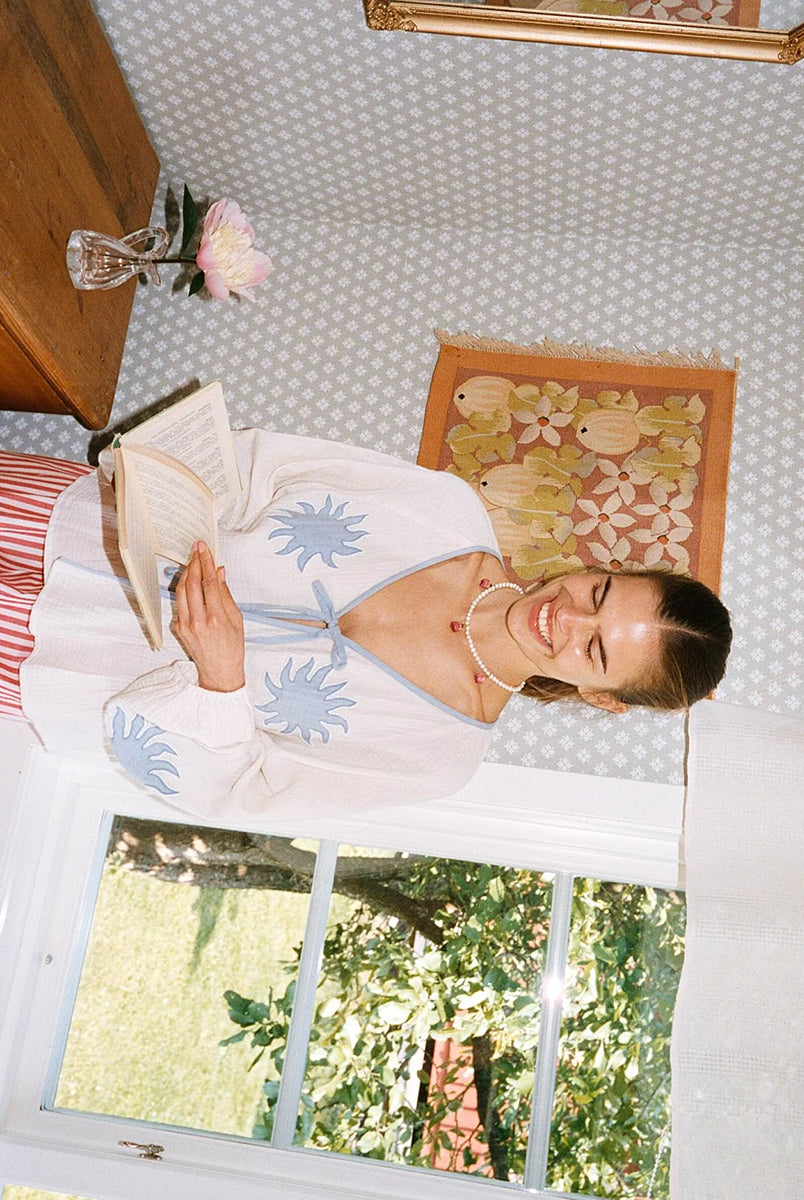 Luźna, kobieca bluzka Ramona Off White marki SISSEL EDELBO wykonana z miękkiej, waflowej tkaniny o podwójnej fakturze, wykonanej z bawełny organicznej. Posiada odcięcie w talii z marszczeniami, które tworzą sylwetkę z falbanką. Ma szerokie rękawy typu raglan z marszczeniami na ramionach i elastycznymi zakończeniami rękawów. Posiada głęboki dekolt w serek i dwa sznurki do wiązania z przodu. Ozdobiony jest haftem z motywem słońca i koralikami z masy perłowej.
-ONE SIZE