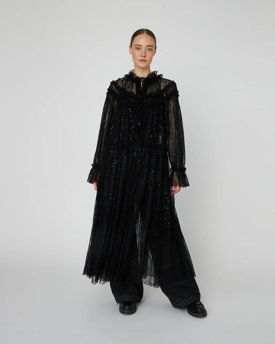 Długa, przezroczysta sukienka Sheer Lace Maxi z koronki z cekinami. Ma zabudowany, efektowny dekolt oraz długie rękawy. Z przodu, na długości ma zapięcia. Można ją nosić zapięta jako sukienkę, bądź rozpiętą jako pelerynę. Sukienka nie posiada podszewki. 