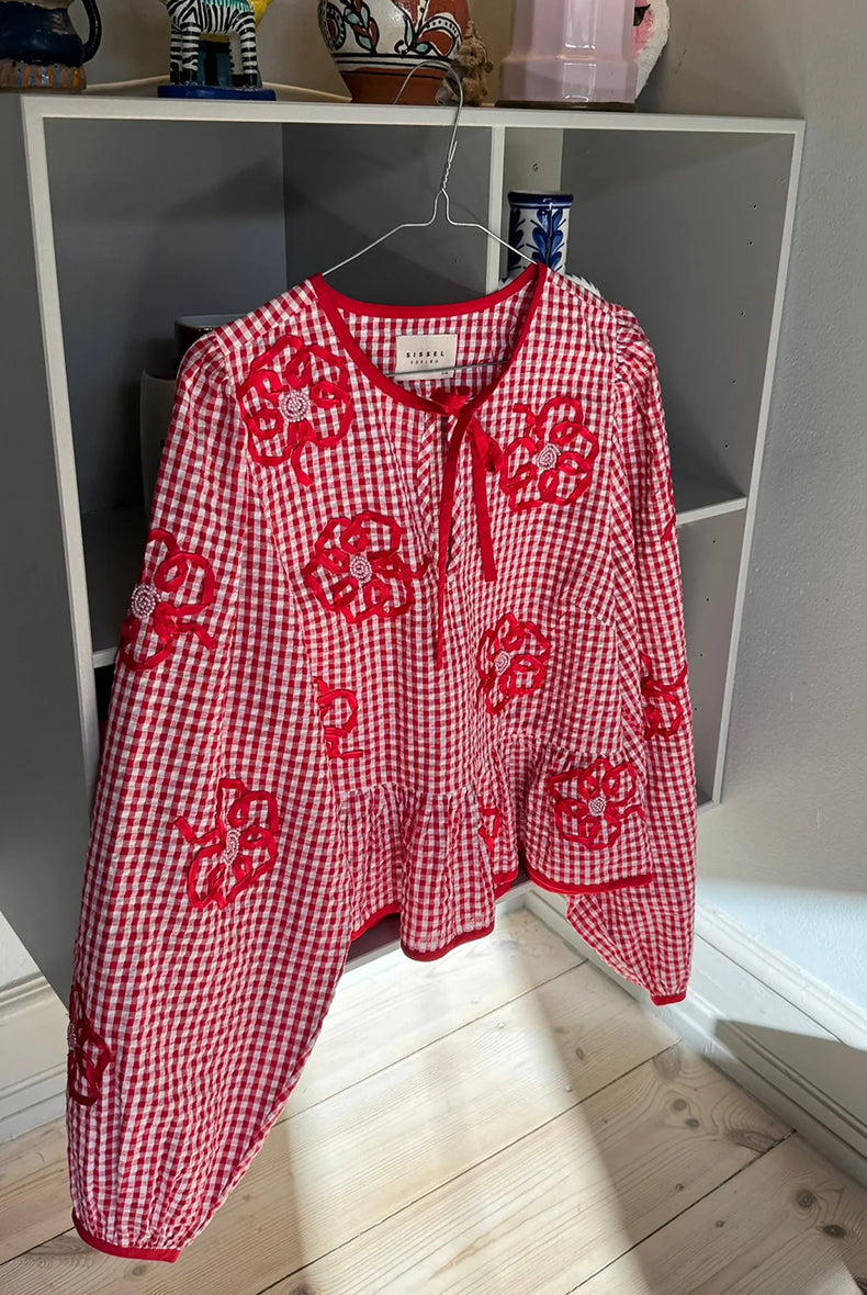 Kobieca bluzka Sunny Red Checks SISSEL EDELBO z szerokimi, bufiastymi rękawami z solidnymi mankietami w kontrastującym kolorze i dekoltem w serek z wiązaniem z przodu. U dołu zakończona falbanką. Ozdobiona jest delikatnymi aplikacjami kwiatowymi z satynowych wstążek i koralików. Wykonana z resztek barwionej przędzy.
-Bufiaste rękawy
-Zakończona falbanką
-Kwiatowe aplikacje z koralikami