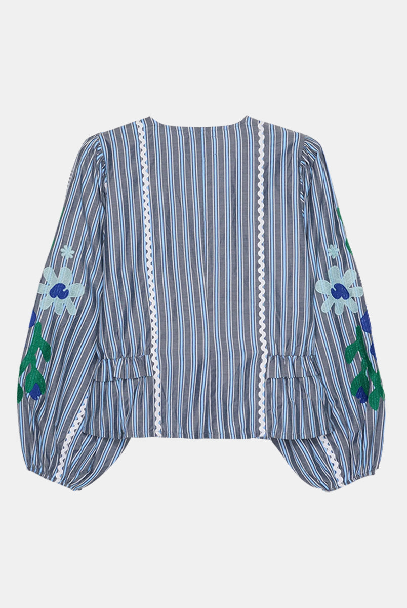 Luźna, koszulowa bluzka Veda Grey Stripes marki SISSEL EDELBO z miękkiej bawełny w szare paski. Ma głęboki dekolt w serek i zapinana jest na ciemnie guziki z masy perłowej z elastycznymi pętelkami. Koszula ma talię z drobnymi falbankami i wstawkami po bokach oraz panele biegnące wzdłuż przodu. Szerokie, bufiaste rękawy zdobi duży wyrazisty haft kwiatowy i zygzakowata bawełniana taśma. Otwory na rękawy posiadają gumkę wewnątrz, co zapewnia wygodne dopasowanie. Uszyta została z bawełnianych resztek tkanin.&nb