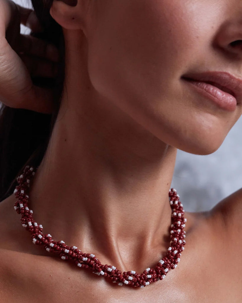 Gruby, efektowny naszyjnik o skręconym splocie Rodeo Anni Lu wykonany drobnych koralików w pięknym, burgundowym kolorze. Wykończony pozłacanym łańcuszkiem o regulowanej długości z niewielką, naturalną perłą na jego końcu.