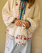 Torebka na ramię Babba Drawstring Off White marki SISSEL EDELBO z bawełny organicznej. Zmieścisz w niej niezbędne rzeczy na co dzień. Ma okrągłe dno i można ją zamknąć za pomocą sznurków. Ozdobiona jest uroczymi, niebieskimi kropkami i haftem krzyżykowym wzdłuż dolnej krawędzi, a także małym, haftowanym napisem SE.