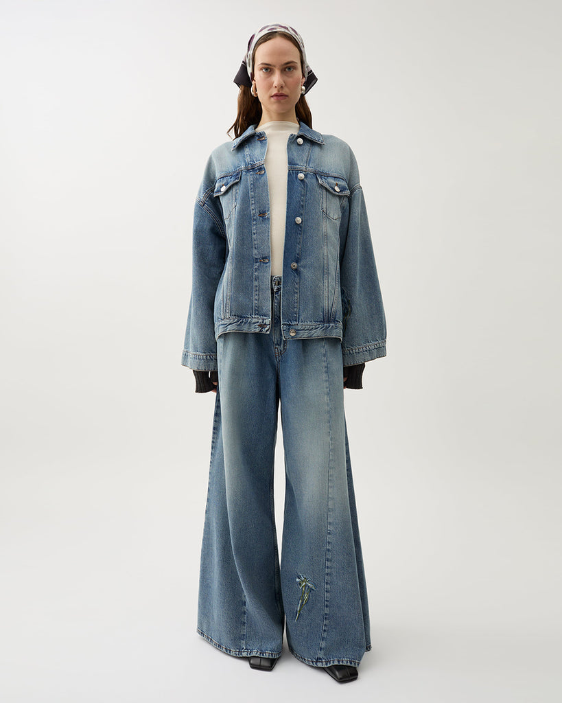 Oversizowa, jeansowa kurtka Bat Indigo w klasycznym, niebieskim odcieniu denimu z kwiatowymi haftami. Posiada lekko rozjaśnione partie na przodzie i rękawach, co tworzy wrażenie vintage. Ma obniżoną linię ramion i szerokie rękawy. Zapięcie na metalowe guziki typowe dla kurtki jeansowej oraz klasyczne kieszenie z patkami.&nbsp;