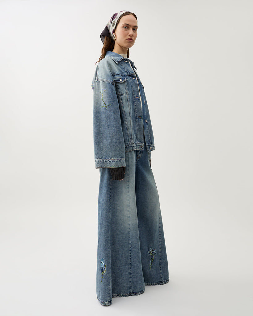 Oversizowa, jeansowa kurtka Bat Indigo w klasycznym, niebieskim odcieniu denimu z kwiatowymi haftami. Posiada lekko rozjaśnione partie na przodzie i rękawach, co tworzy wrażenie vintage. Ma obniżoną linię ramion i szerokie rękawy. Zapięcie na metalowe guziki typowe dla kurtki jeansowej oraz klasyczne kieszenie z patkami.&nbsp;