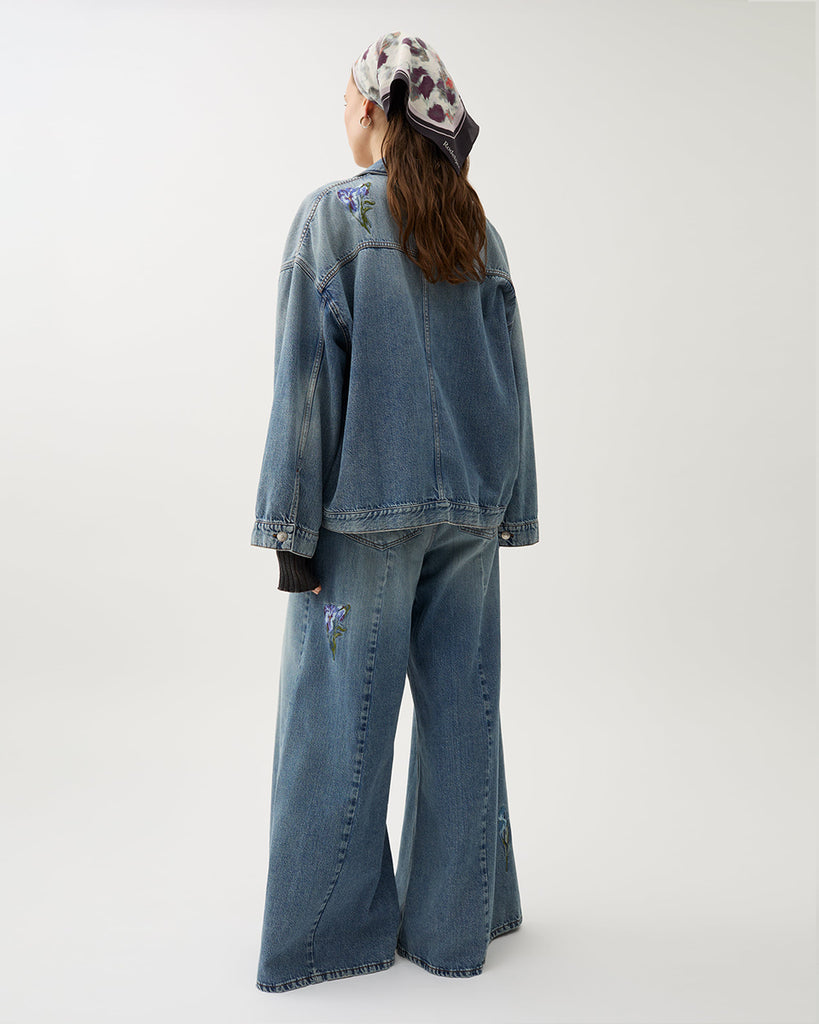 Oversizowa, jeansowa kurtka Bat Indigo w klasycznym, niebieskim odcieniu denimu z kwiatowymi haftami. Posiada lekko rozjaśnione partie na przodzie i rękawach, co tworzy wrażenie vintage. Ma obniżoną linię ramion i szerokie rękawy. Zapięcie na metalowe guziki typowe dla kurtki jeansowej oraz klasyczne kieszenie z patkami.&nbsp;