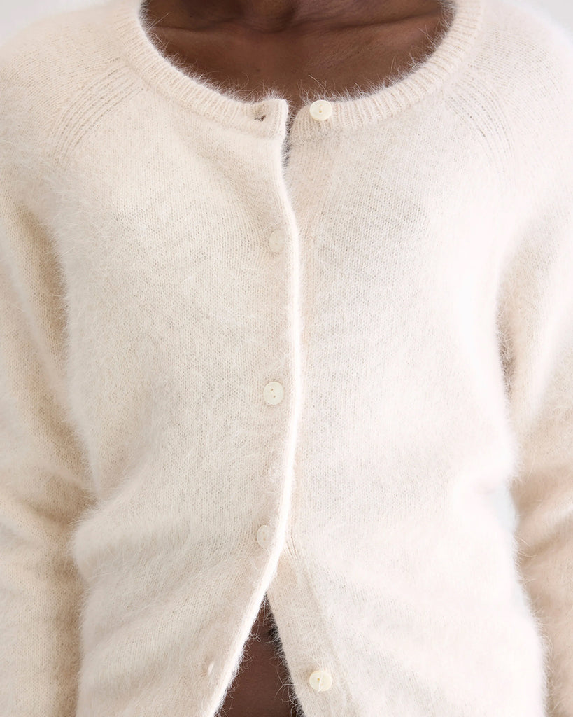 Prosty, krótki, kremowy kardigan Datair Bellerose z okrągłym dekoltem. Wykończony ściągaczami. Zapinany na guziki. Sweter ma prosty krój i wykonany jest z bardzo miękkiej i puchatej dzianiny z angorą