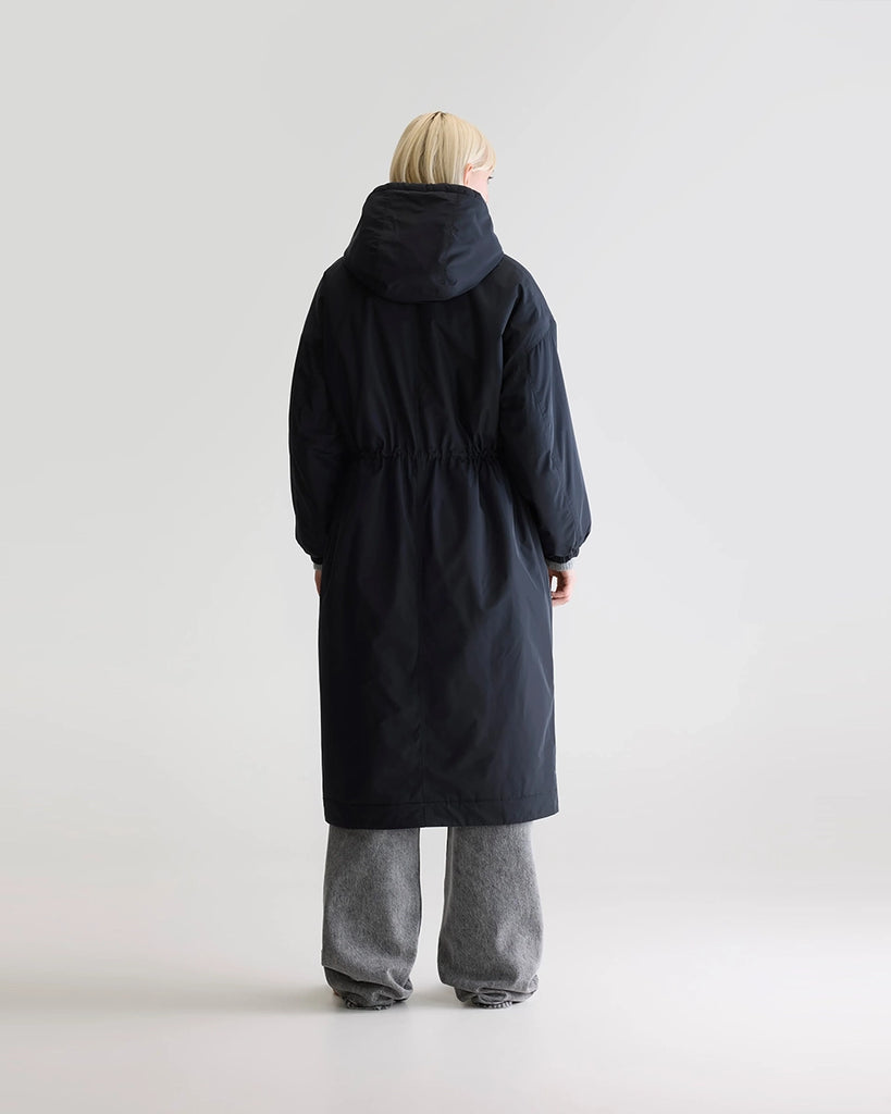 Długi, granatowy płaszcz Hosfo Bellerose z kapturem, wysokim kołnierzem podszyty sztucznym futrem w kontrastowym kolorze. Jednocześnie kobiecy i praktyczny. Zwężany i wiązany w talii. Wykonany z tkaniny z powłoką przeciwdeszczową. Płaszcz ma dwie, boczne kieszenie, rękawy wykończone ściągaczami i długość za kolana