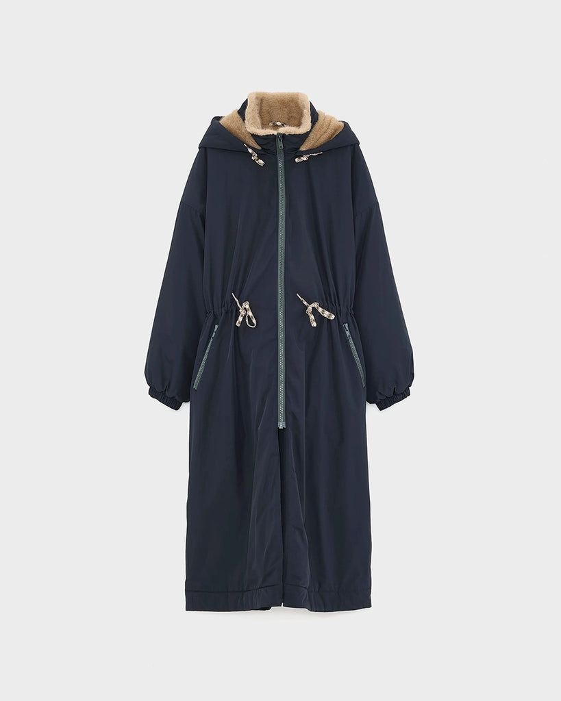 Długi, granatowy płaszcz Hosfo Bellerose z kapturem, wysokim kołnierzem podszyty sztucznym futrem w kontrastowym kolorze. Jednocześnie kobiecy i praktyczny. Zwężany i wiązany w talii. Wykonany z tkaniny z powłoką przeciwdeszczową. Płaszcz ma dwie, boczne kieszenie, rękawy wykończone ściągaczami i długość za kolana