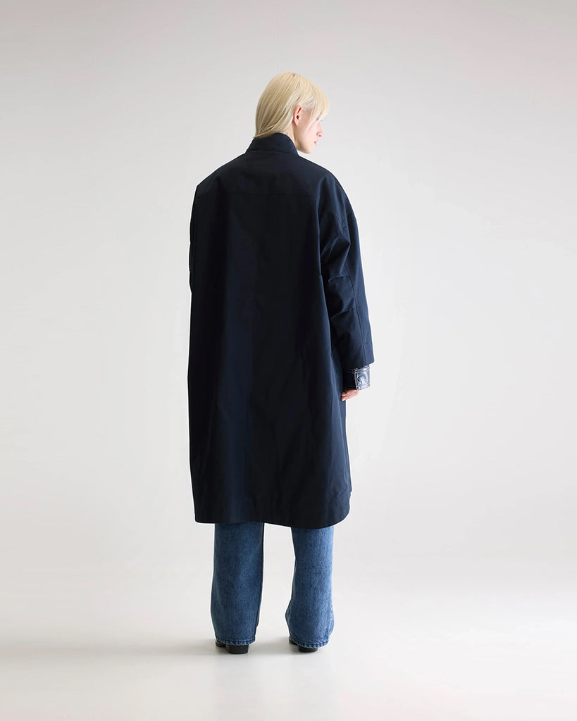 Długi, oversizowy, przejściowy trencz Juttac Bellerose z wysokim kołnierzem, opadającymi ramionami i luźnymi rękawami. Zapinany na jeden, boczny rząd guzików. Płaszcz ma prosty krój i głębokie, boczne rozcięcia zapinane na guziki. Posiada czerwoną podszewkę w kratę. 